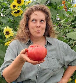 Christy-Anderson-Mt-Gile’s-Famous a farmer in front of sunflowers holds a large red heirloom tomato in one hand while looking playfully to one side