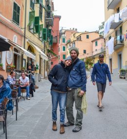 Nomad Pasta in Italy Two smiling people stand on a busy village street in Italy; they are flanked by pedestrians and people eating at an outdoor cafe and colored buildings rise behind them